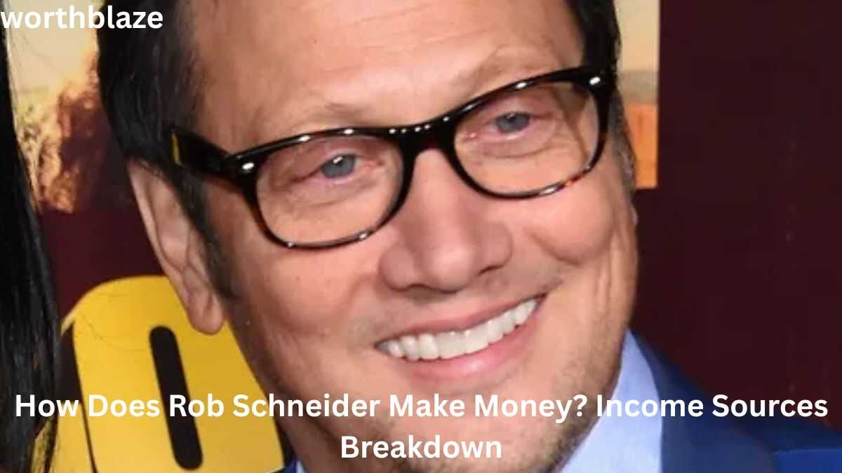 Rob Schneider smiling at a public event, wearing glasses and a blue suit, featured in an image discussing income sources and Rob Schneider Net Worth.