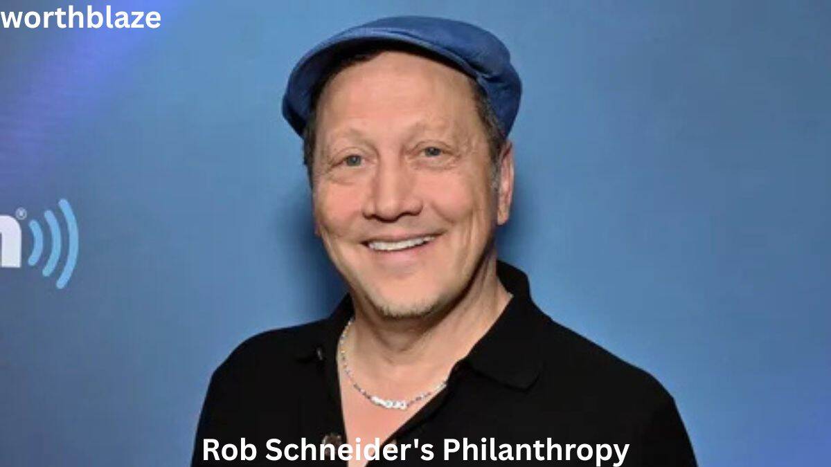 Rob Schneider smiling in front of a blue backdrop while discussing charitable work and community involvement, featured in a Rob Schneider Net Worth article.